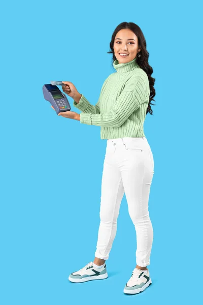 Young Asian woman with payment terminal and credit card on blue background