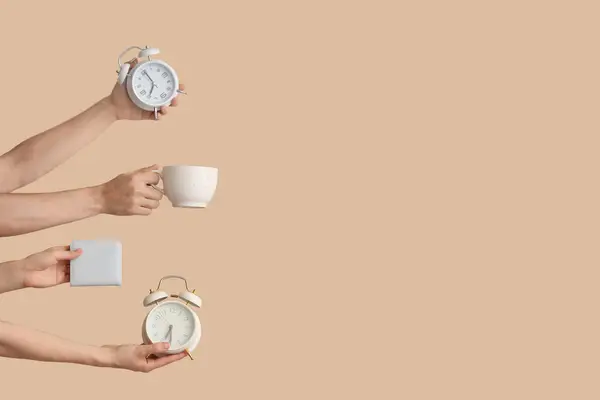 Hands holding alarm clocks, cup and wallet on beige background