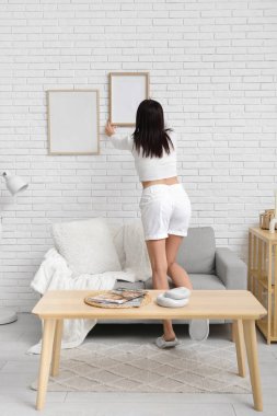 Oturma odasındaki beyaz tuğla duvara boş bir çerçeve asan genç kadın.