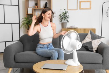 Evde elektrikli vantilatörü olan genç bir kadın.