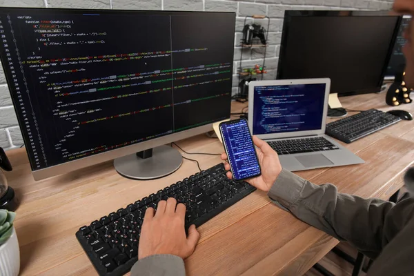 Male programmer working with mobile phone and computer at table in office, closeup