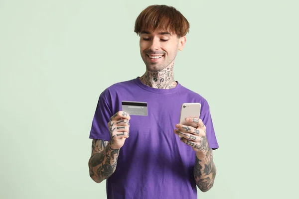 Tattooed young man with credit card using mobile phone on green background