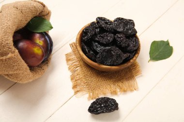 Bir kase lezzetli kurutulmuş erik ve beyaz ahşap arka planda taze meyve dolu çuval.