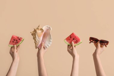 Olgun karpuz dilimleri, güneş gözlüğü ve bej arka planda deniz kabuğu olan kadın elleri.