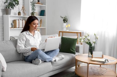 Oturma odasındaki kanepede dizüstü bilgisayarlı güzel bir Asyalı kadın oturuyor.