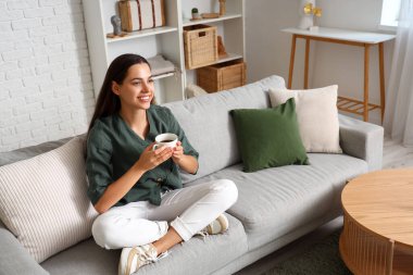 Oturma odasındaki kanepede oturan bir fincan kahveyle güzel genç bir kadın.