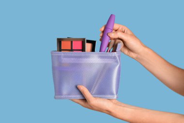 Mavi arka planda dekoratif kozmetik ürünleri olan bir çanta tutan kadın.