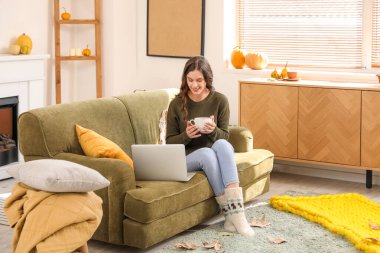 Bir fincan çay ve dizüstü bilgisayarı olan genç bir kadın sonbahar günü evde oturuyor.
