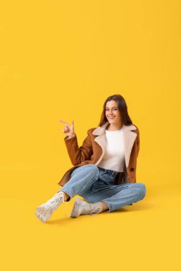 Kışlık elbiseli genç kadın sarı arka plandaki bir şeyi işaret ediyor.