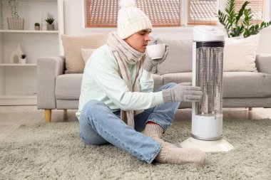 Evde elektrikli ısıtıcının yanında çay ısıtan genç bir adam.