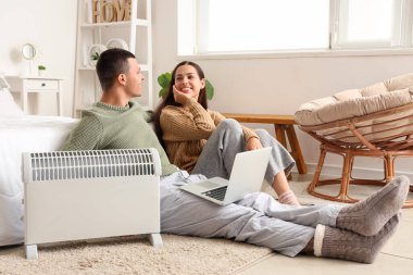 Yatak odasındaki radyatörün yanında laptoplu genç çift ısınıyor.