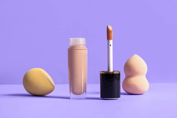 Bottle of makeup foundation and sponges on purple background