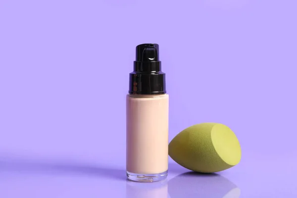 Bottle of makeup foundation and sponge on purple background