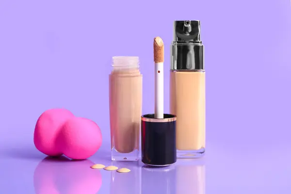 Bottles of makeup foundation and sponge on purple background