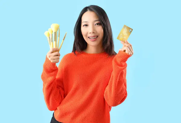 青い背景にクレジットカードとメイクブラシを持つ若いアジアの女性 — ストック写真