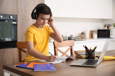 Kulaklık ve dizüstü bilgisayarı olan küçük çocuk mutfakta internetten ders çalışıyor.