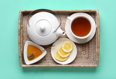 Çaydanlıklı hasır tepsi, bir fincan çay, bal ve arkaplanda limon dilimleri.