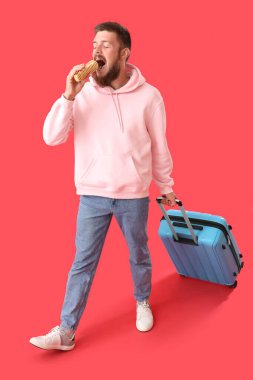 Kırmızı arka planda lezzetli sosisli yiyen bavullu yakışıklı genç adamın portresi.