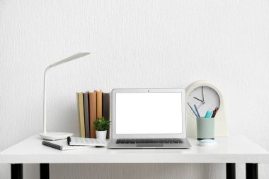 Beyaz duvarın yanındaki masada defter ve lamba olan modern bir laptop.