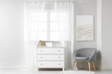 Açık renkli oturma odası, pencerenin yanındaki gri koltuk ve çekmeceler.