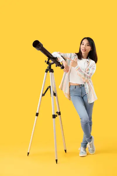 Young Asian woman with telescope on yellow background