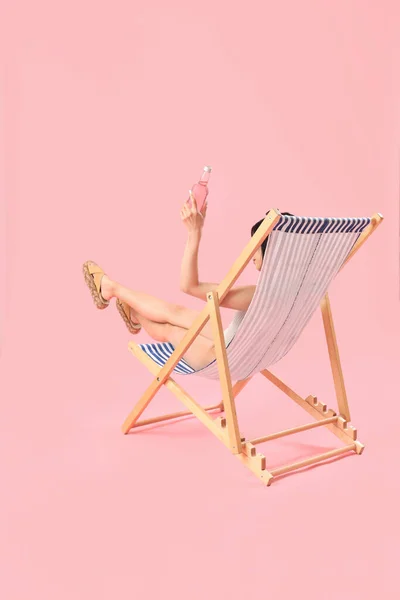 Beautiful Asian woman with soda relaxing in deck chair on pink background
