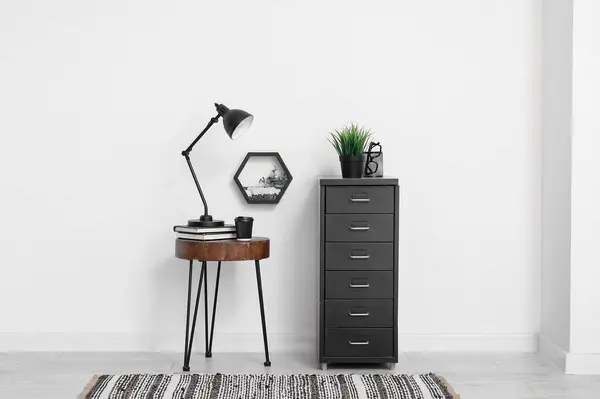 Desk lamp on small table and file cabinet with houseplant near white wall