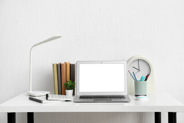 Modern laptop with notebooks and lamp on table near white wall