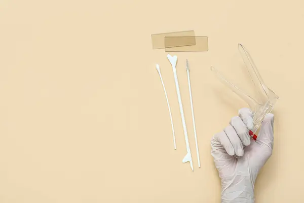 Hand in medical glove, with gynecological speculum and pap smear test tools on beige background