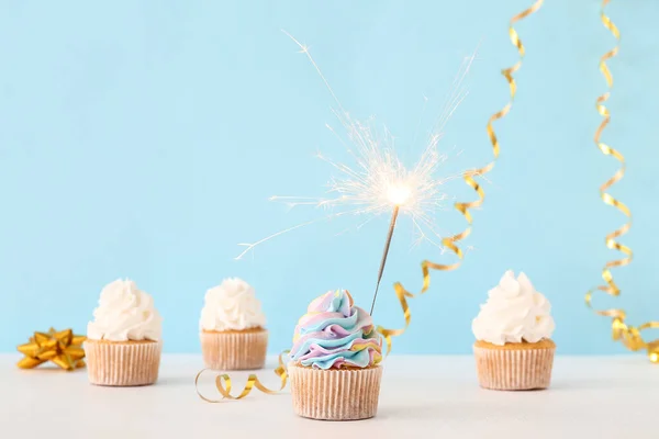 Tasty birthday cupcakes with sparkler and decor on blue background
