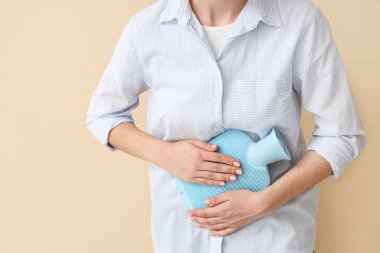 Bej arka planda karın ağrısı çeken, sıcak su şişesi olan genç bir kadın, yakın plan.