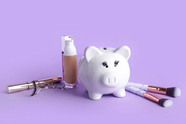 Piggy bank with eyelashes and cosmetic products on lilac background