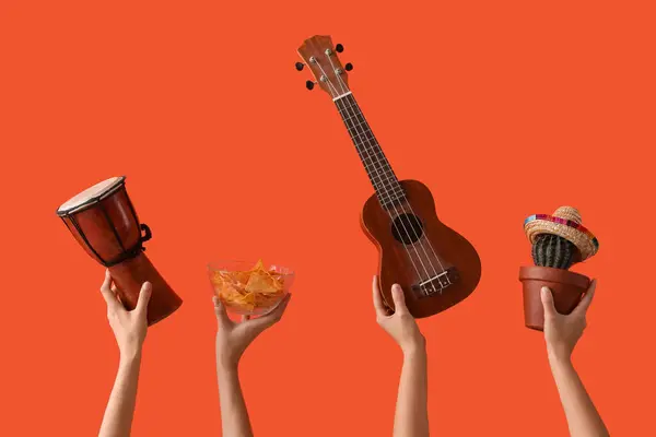 Female hands holding drum, ukulele, nachos and cactus on orange background
