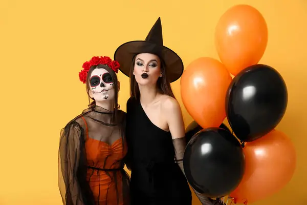 Young women dressed for Halloween with balloons blowing kisses on yellow background