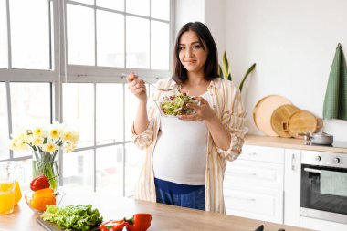 Genç hamile kadın mutfakta sebze salatası yiyor.