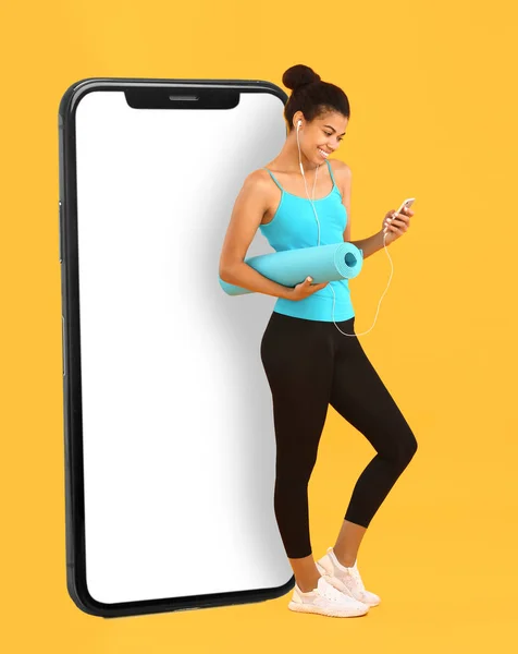 Sporty African-American woman with yoga mat and big smartphone on yellow background. Weight loss concept
