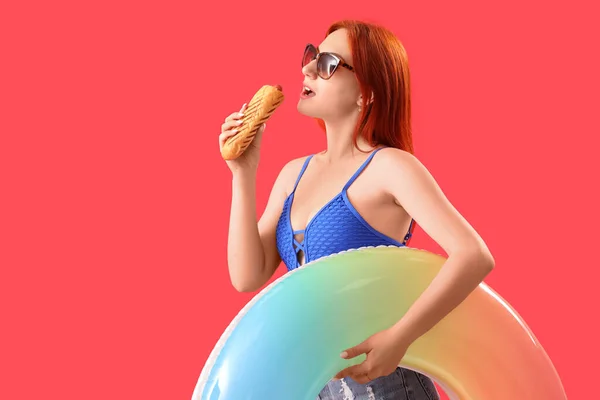 Beautiful young woman with french hot dog and inflatable ring on red background