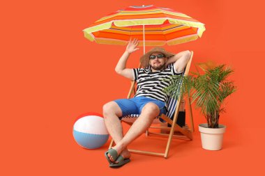 Şemsiyeli ve şişme toplu yakışıklı genç adam kırmızı arka planda şezlongda oturuyor.