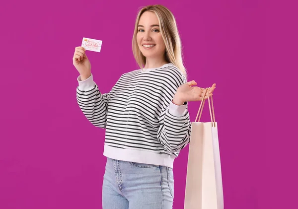 Young woman with gift card and shopping bag on purple background