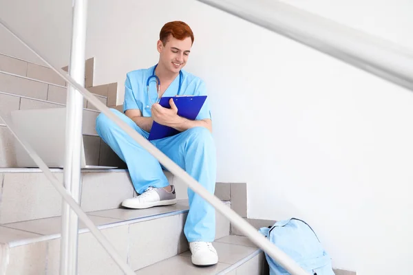 Male medical student with clipboard sitting on stairs at university