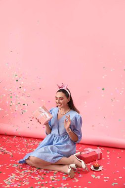 Doğum günü hediyeli mutlu genç kadın, pembe arka planda kek ve konfeti.