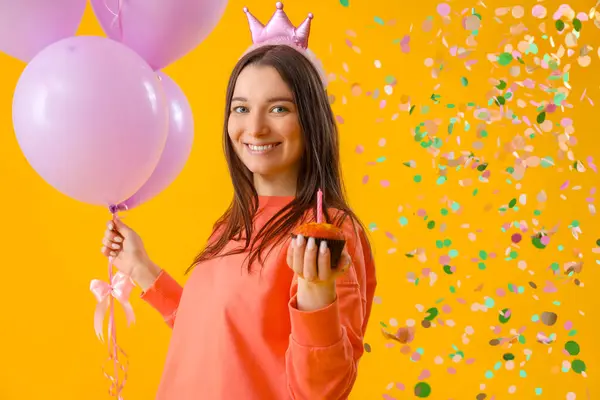 Happy young woman with Birthday muffin and balloons on yellow background