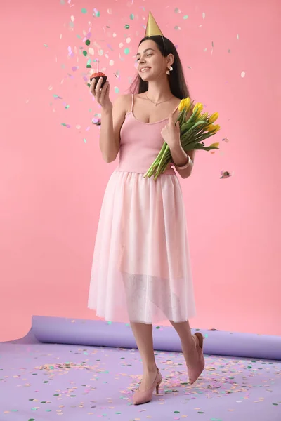 Happy young woman with Birthday muffin and tulips on color background