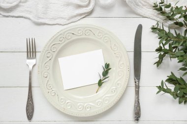 Çatal bıçak takımı, boş kart ve beyaz ahşap arka planda bitki dalları.