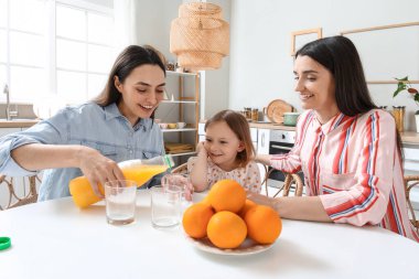 Evlatlık küçük kızı olan genç lezbiyen çift mutfakta portakal suyu içiyor.