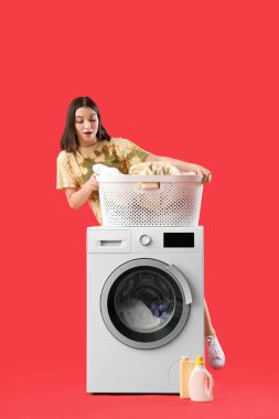 Kırmızı arka planda çamaşır sepeti ve çamaşır makinesi olan genç bir kadın.