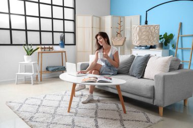 Oturma odasında dizüstü bilgisayar kullanan çay içen genç bir kadın.