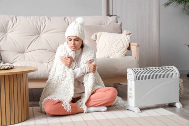 Frozen young woman with plaid warming near radiator at home