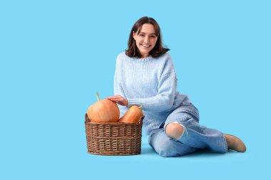 Mavi arka planda oturan sepet dolusu balkabağı olan genç kadın.