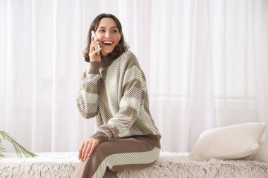 Sıcak kazaklı genç bir kadın evde cep telefonuyla konuşuyor.